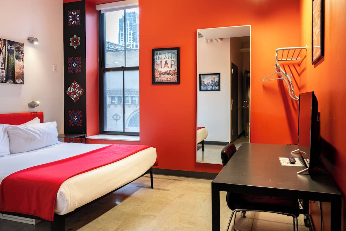 A hotel room with a bed, a desk, a chair, a window with a city view, and a mirror on the wall.