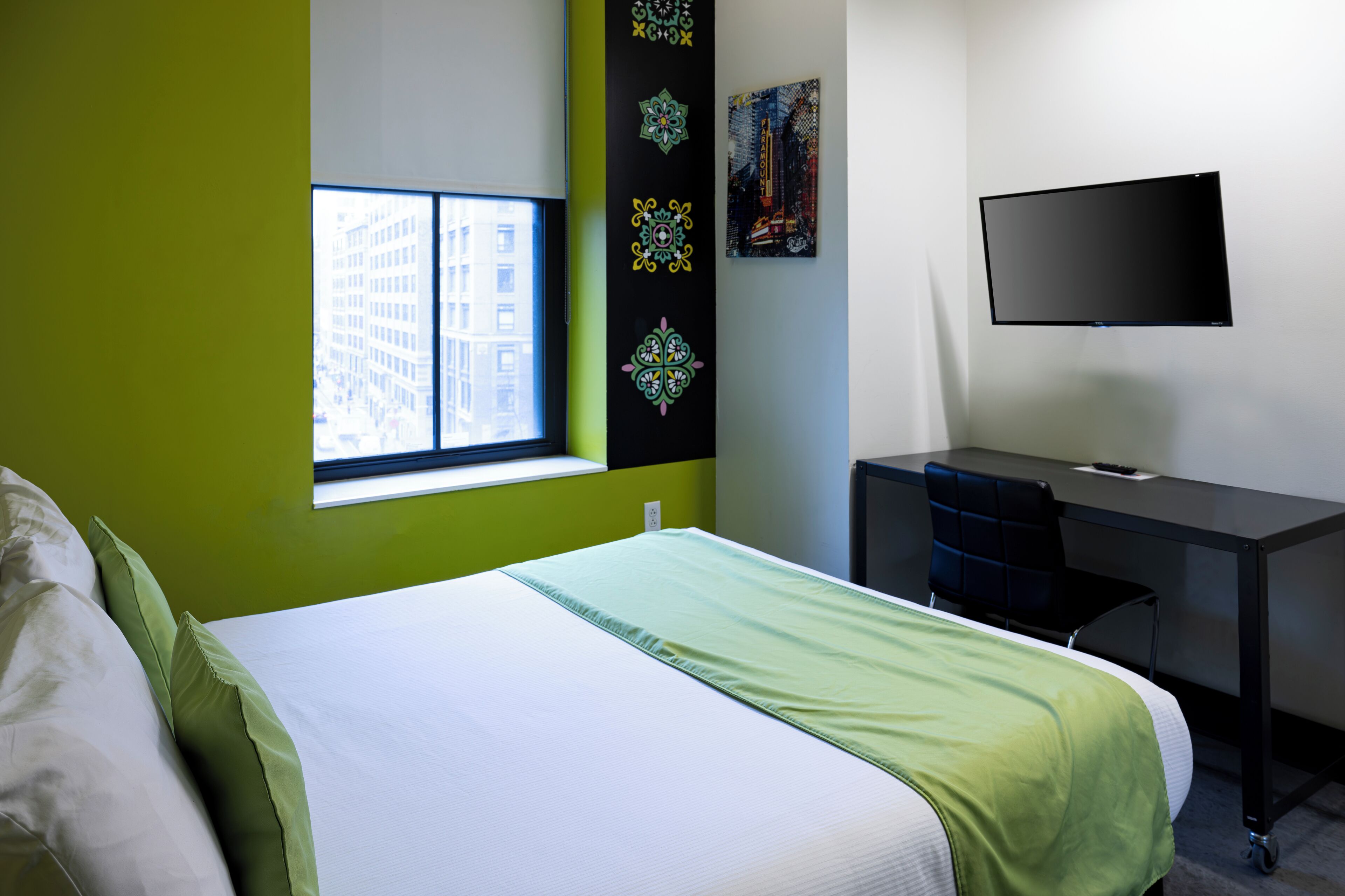Quarto de hotel com cama, escrivaninha, televisão e janela vista para a cidade.
