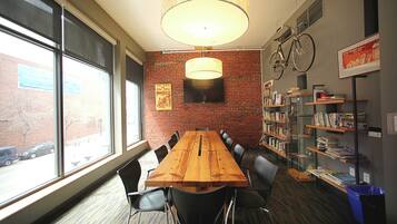 A conference room with a long wooden table, black chairs, a brick wall, a mounted TV, and a bookshelf with books and a bicycle.