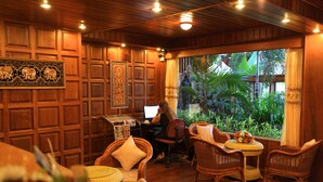 A room with wooden paneling, wicker furniture, a computer desk, and a view of tropical plants. - The Hotel Emperor (Mandalay)