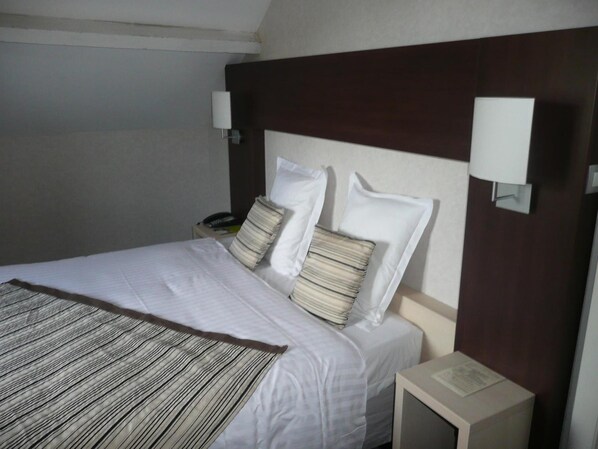 A hotel room with a neatly made bed, two bedside lamps, a nightstand with a phone, and a small table with a menu or information card.