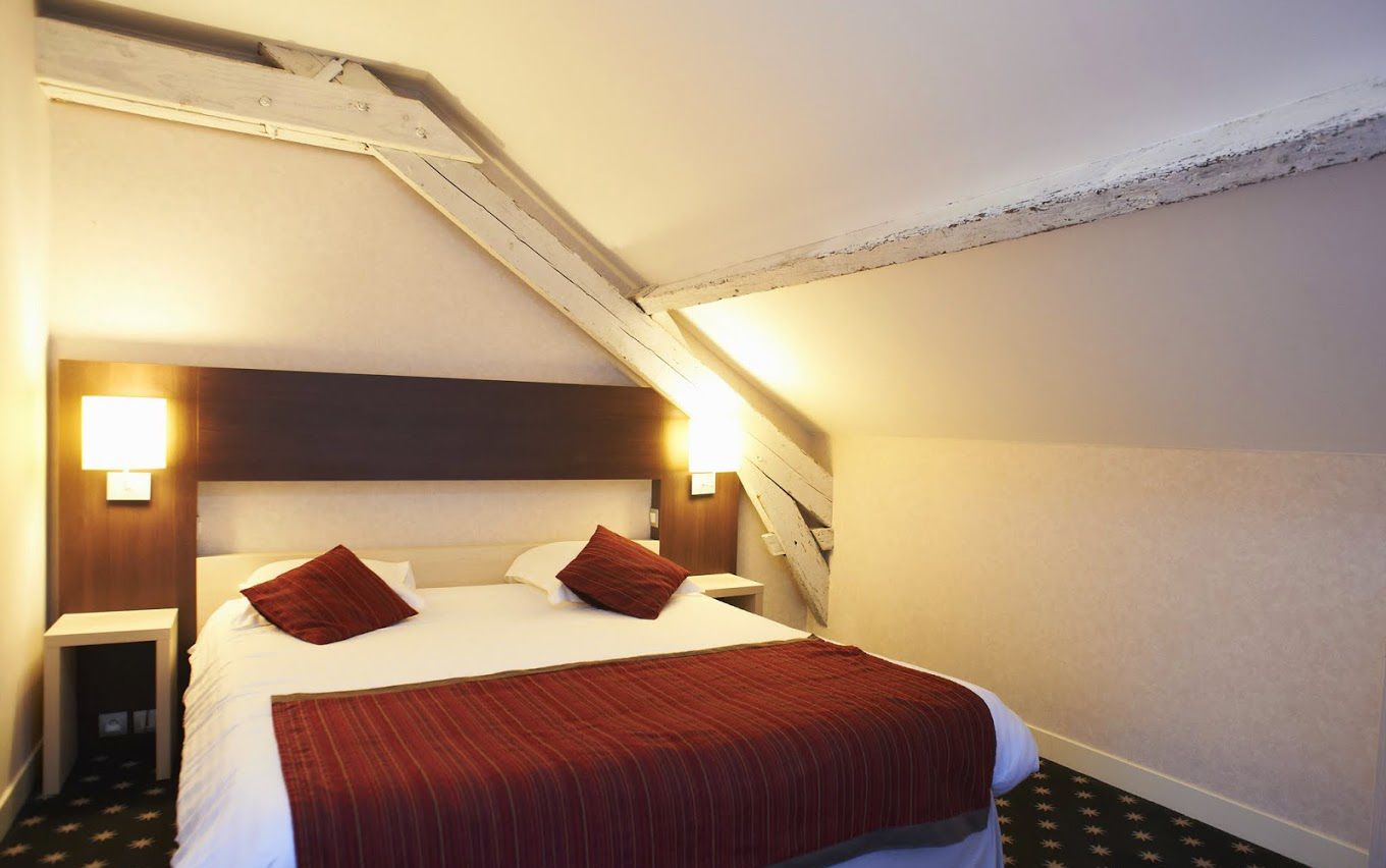 Habitación de hotel con cama, dos lámparas de noche, cabecero de madera y techo inclinado.