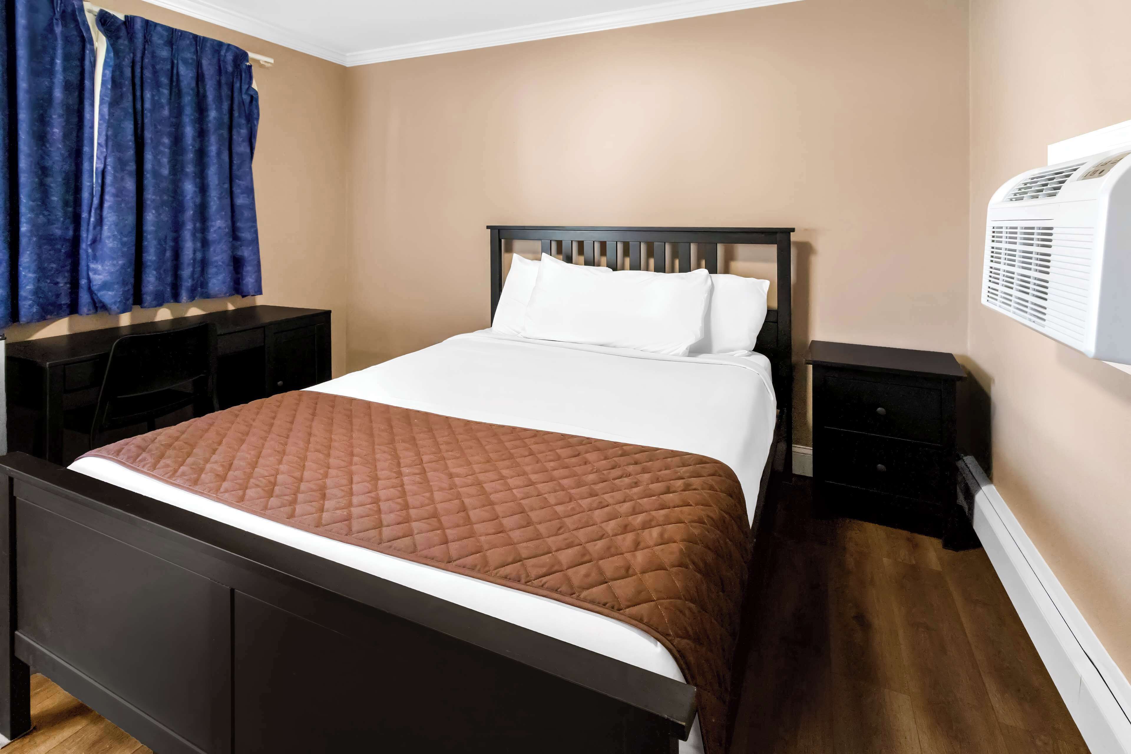 A neatly made bed with a brown quilt, a dark wooden headboard, and white pillows. There are two bedside tables, one with a lamp and the other with a small drawer. A blue curtain is partially drawn, and an air conditioning unit is mounted on the wall.