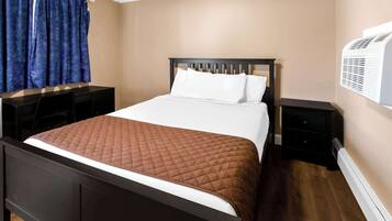 A neatly made bed with a brown quilt, a dark wooden headboard, and white pillows. There are two bedside tables, one with a lamp and the other with a small drawer. A blue curtain is partially drawn, and an air conditioning unit is mounted on the wall.