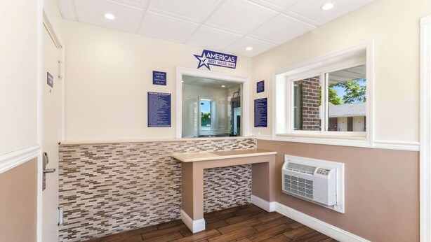 A hotel reception area with a tiled wall, a reception desk, a window with a view, and a wall-mounted air conditioning unit. - Americas Best Value Inn Avenel Woodbridge (Avenel)
