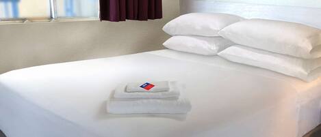Una cama bien hecha con sábanas y almohadas blancas, una toalla doblada y una ventana con cortinas.