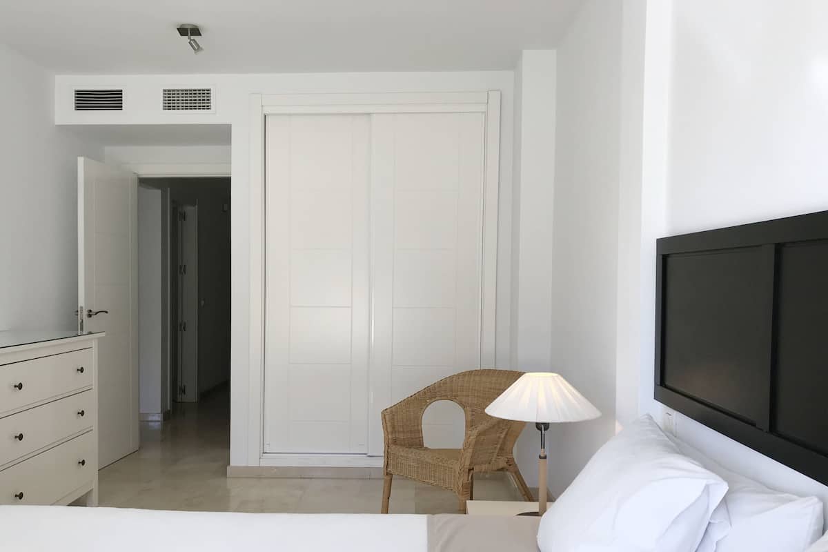 A bedroom with a bed, a dresser, a wicker chair, a lamp, and a closed door.