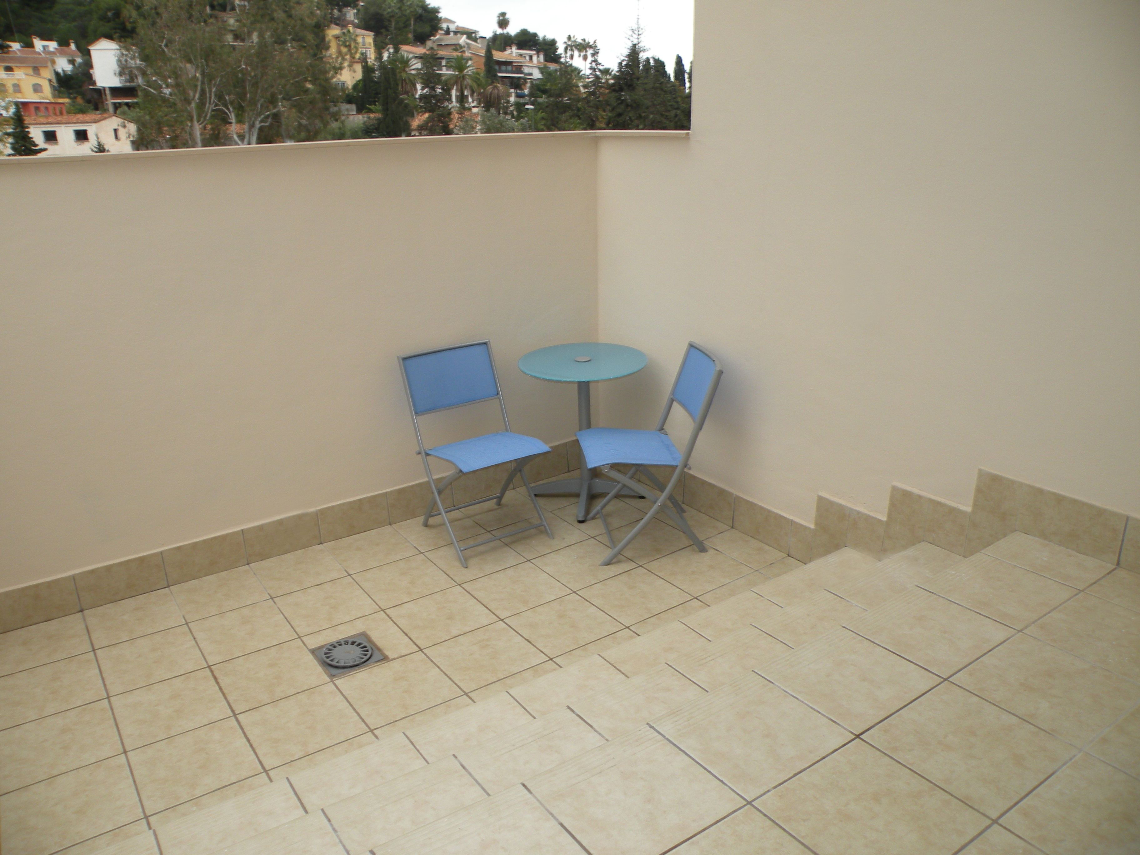 A small outdoor patio with two blue chairs and a round table, surrounded by beige tiles and a low wall.