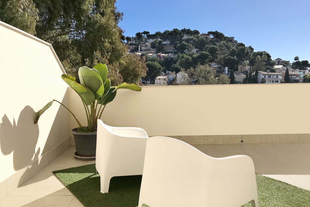 A rooftop terrace with two white chairs, a potted plant, and a view of a hillside town.