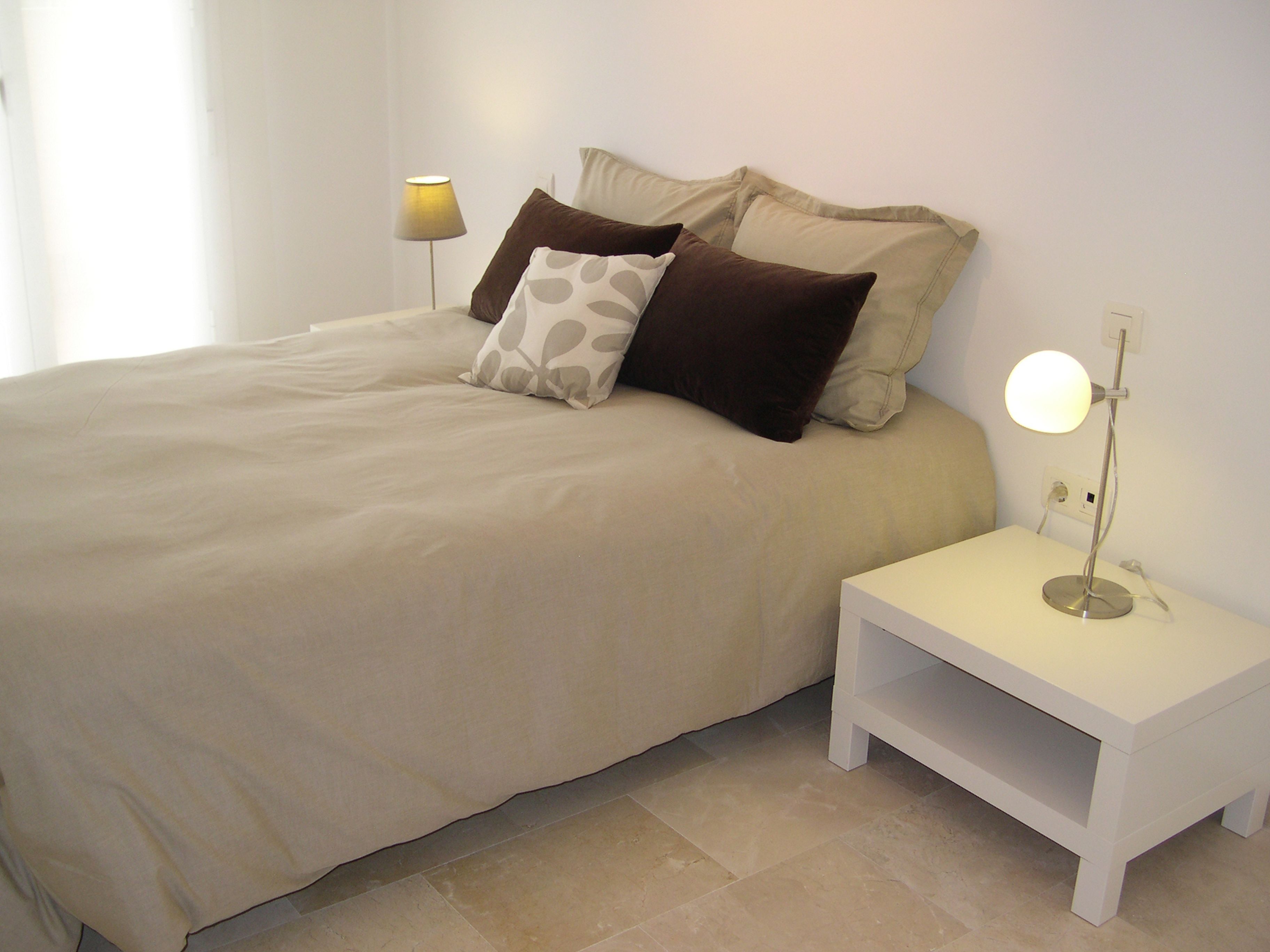 Een opgemaakt bed met een beige sprei en twee kussens, een nachtkastje met een lamp en een witte nachtkastje.