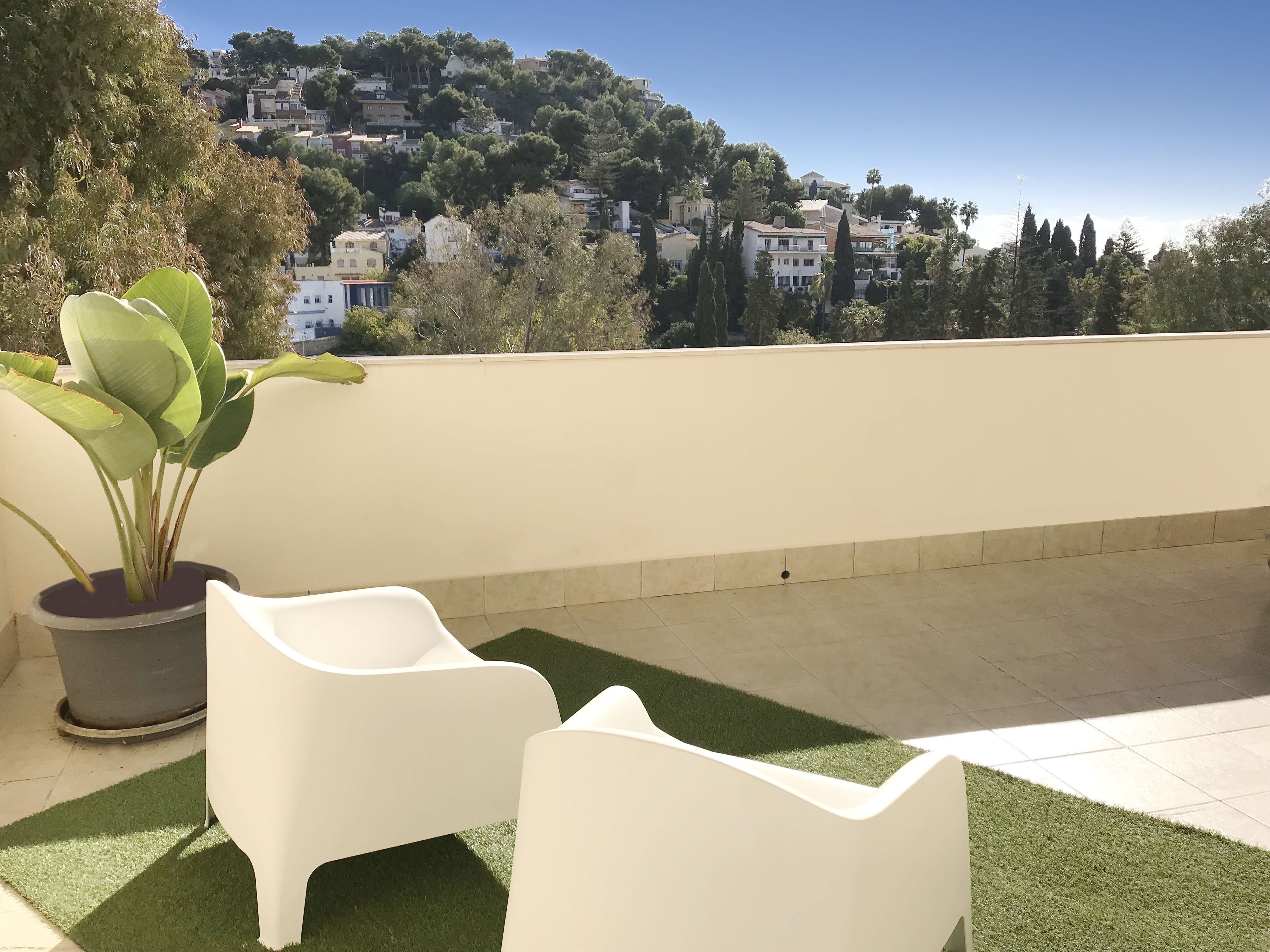 A rooftop terrace with a potted plant, two white chairs, and a view of a hillside with buildings.