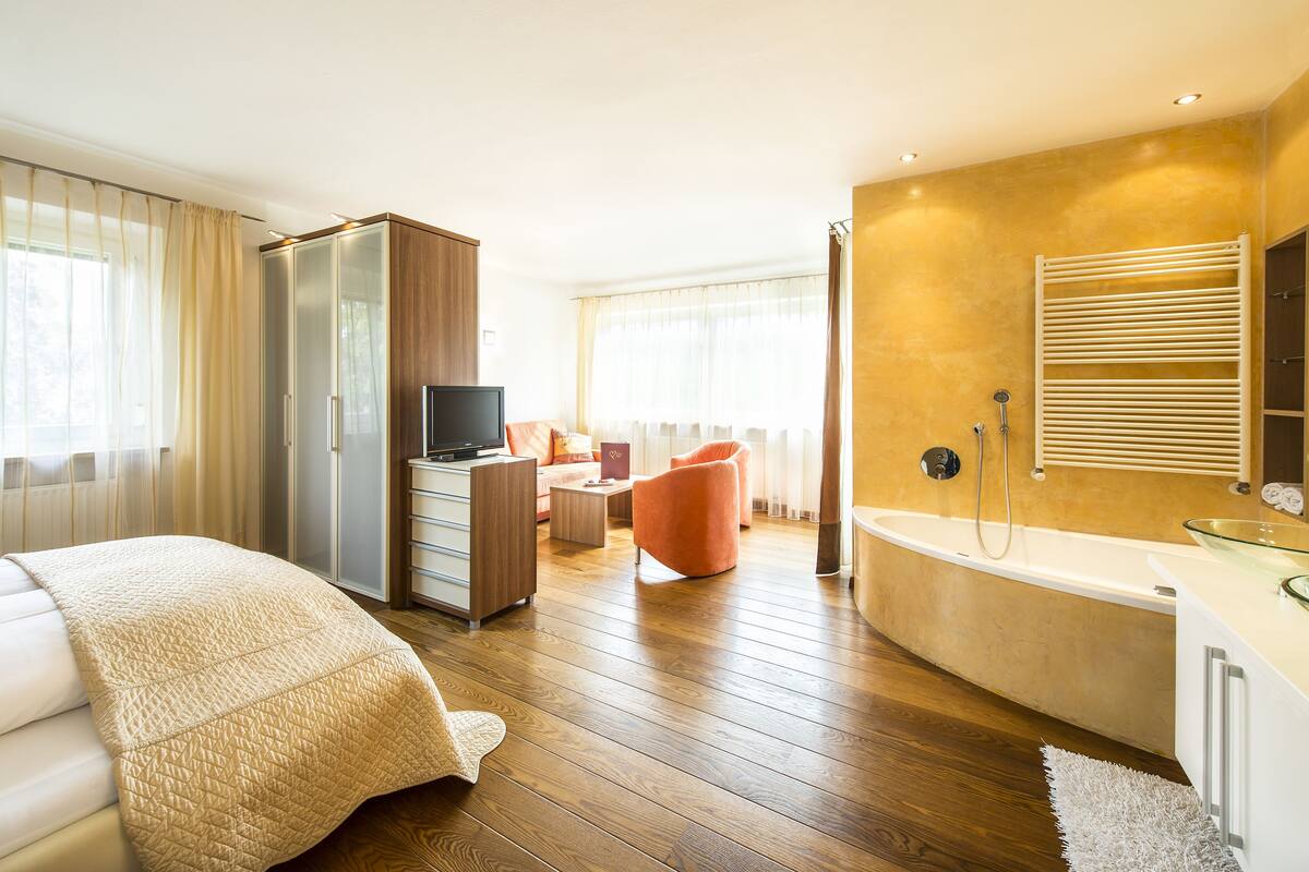 A modern hotel room with a wooden floor, a large bed with a beige quilt, a desk with a laptop, a TV, a sofa, and a bathroom with a bathtub and shower.