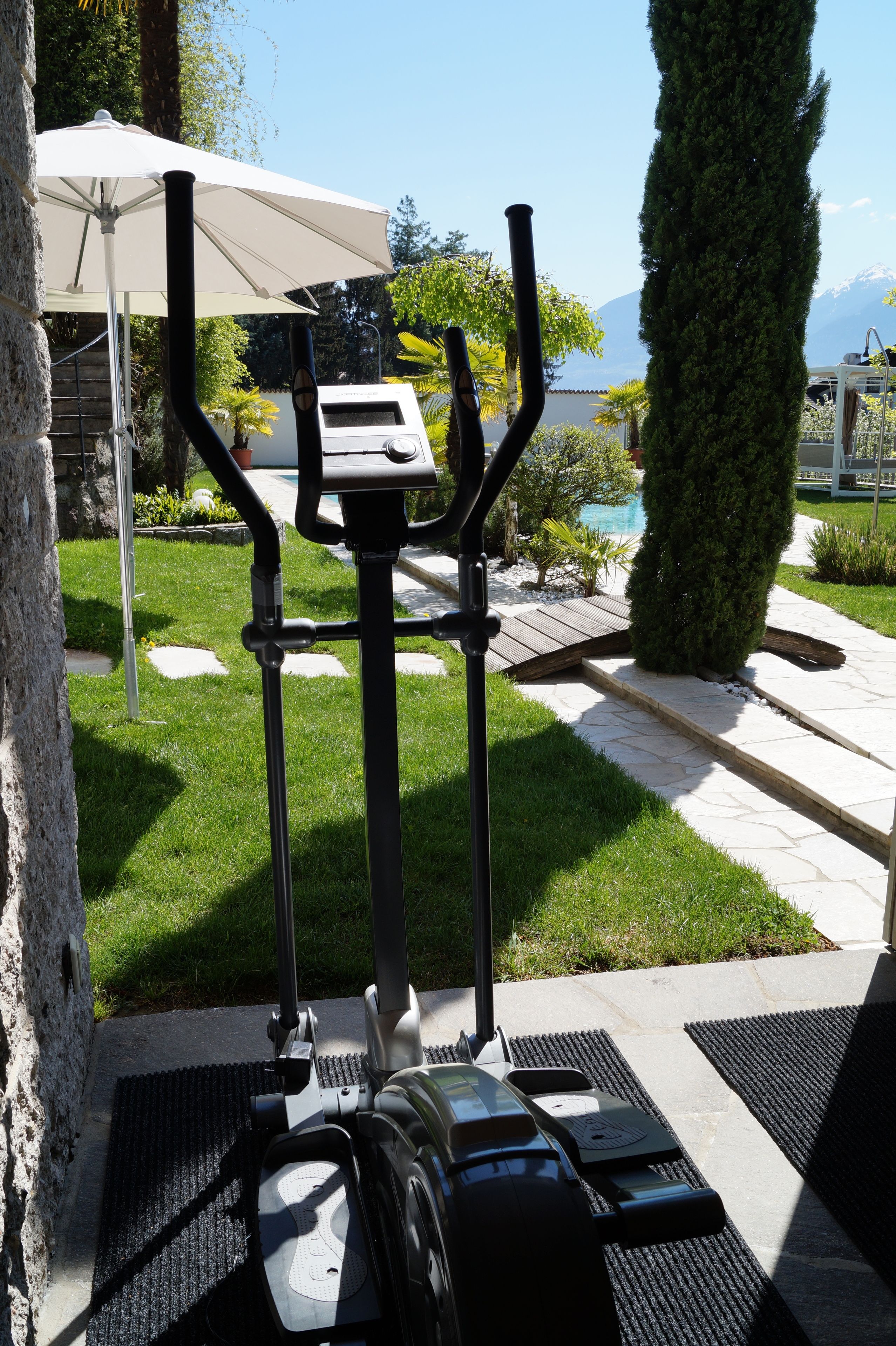 An outdoor exercise machine with a stepper and a stationary bike, positioned on a patio with a pool and greenery in the background.
