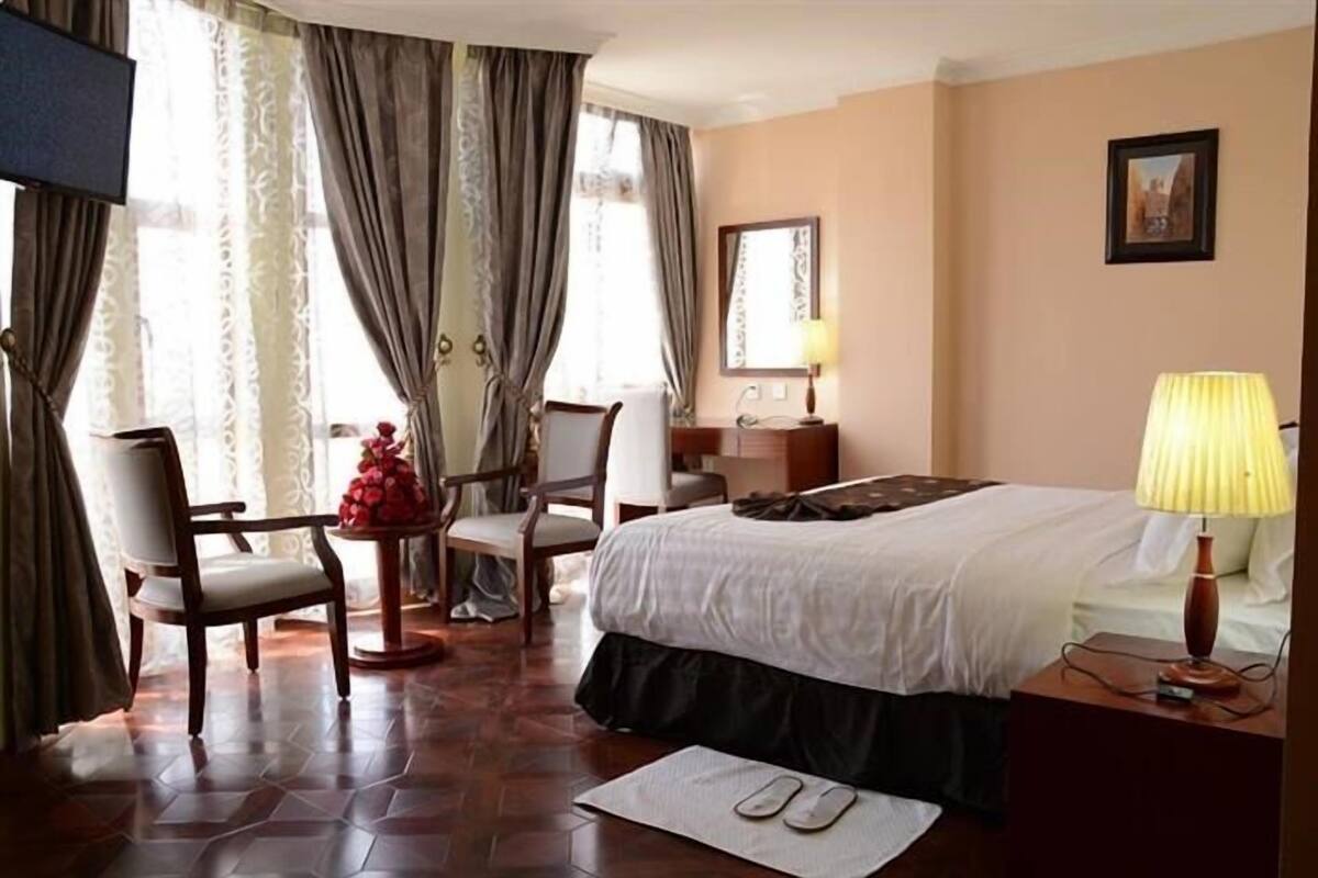 A hotel room with a bed, chairs, a small table with a vase, a lamp, and a mirror on the wall.