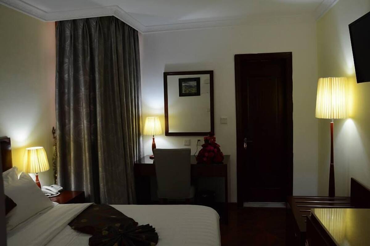 A hotel room with a bed, two bedside lamps, a desk with a chair, a mirror, and a door.
