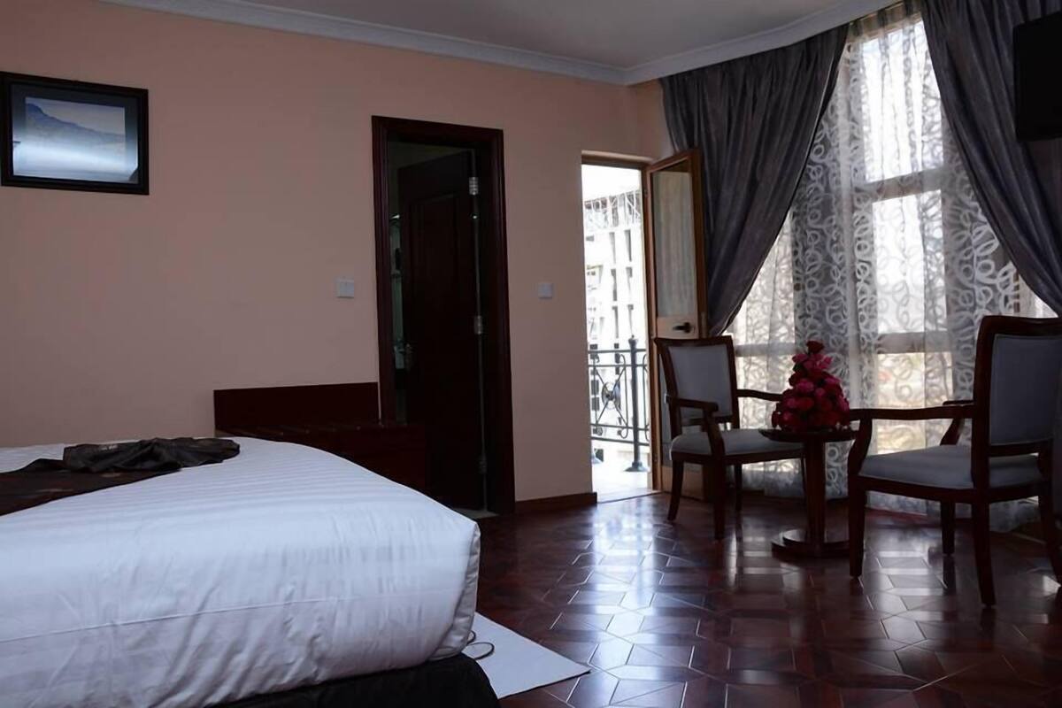 A hotel room with a bed, a dining table with chairs, a balcony with a railing, and a window with curtains.