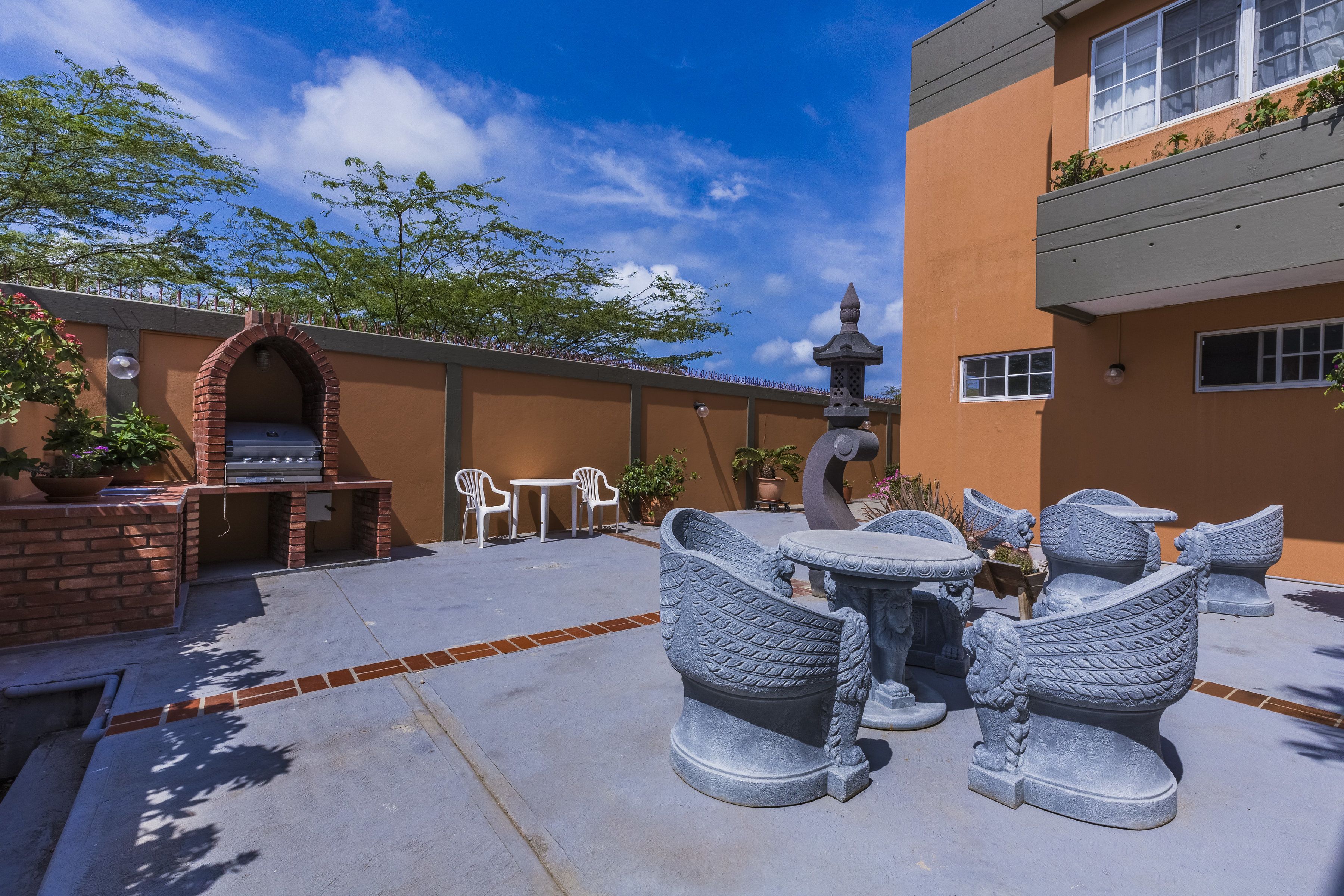An outdoor patio with a brick barbecue, white plastic chairs, and decorative stone furniture.