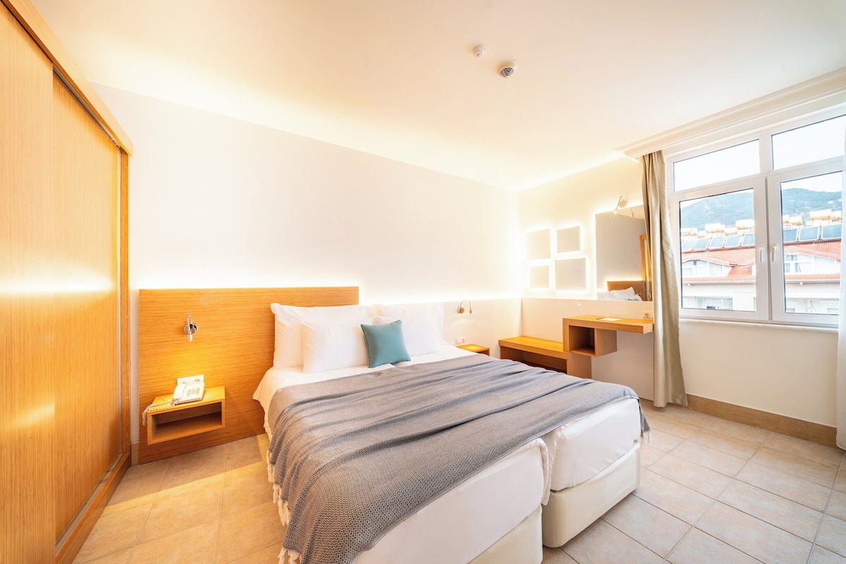 Ett hotellrum med en säng, sängbord, en spegel, ett fönster med utsikt och en garderob i trä.