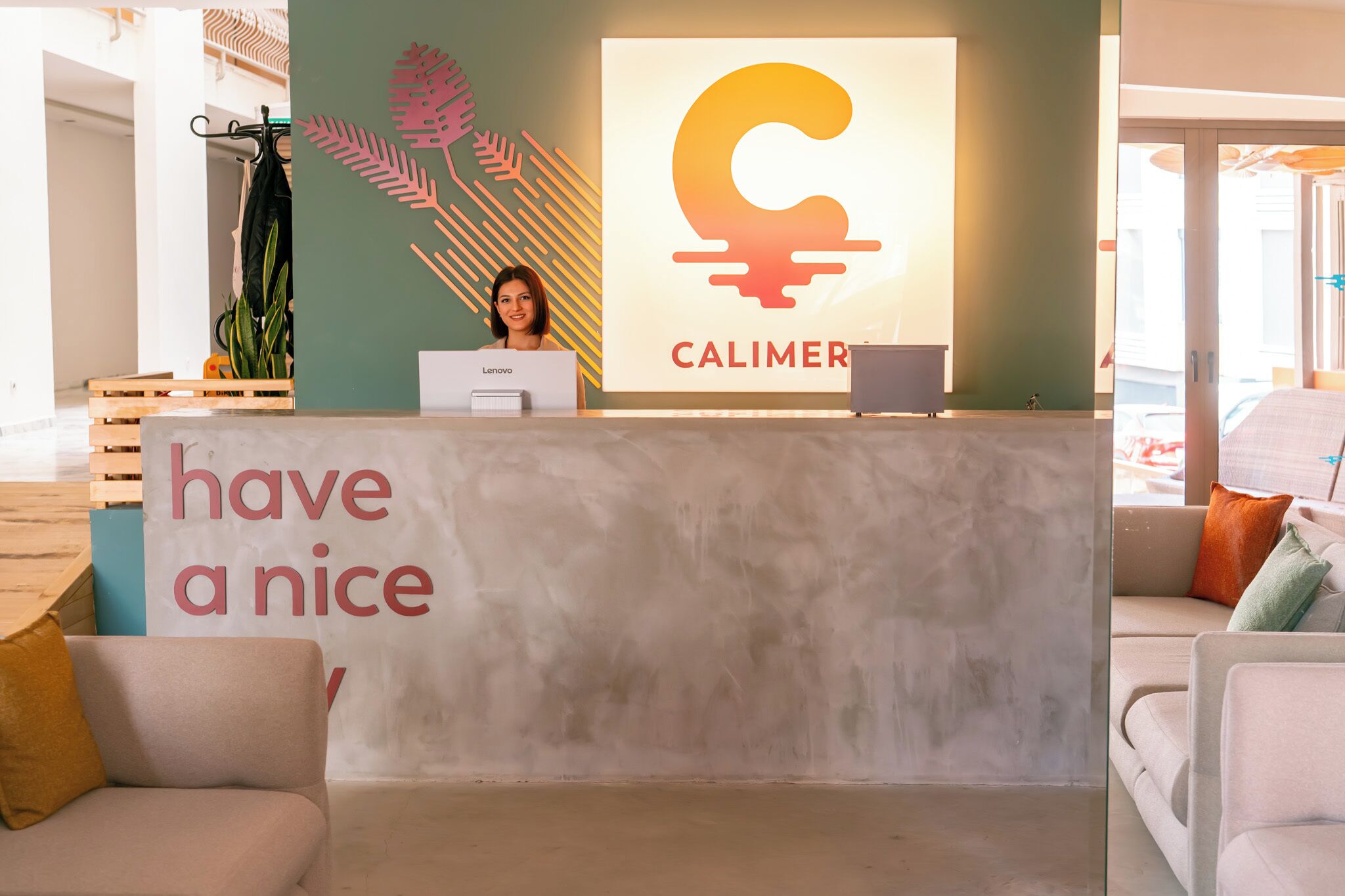A reception desk with a 'have a nice day' sign, a Lenovo computer, and a Calimero logo.