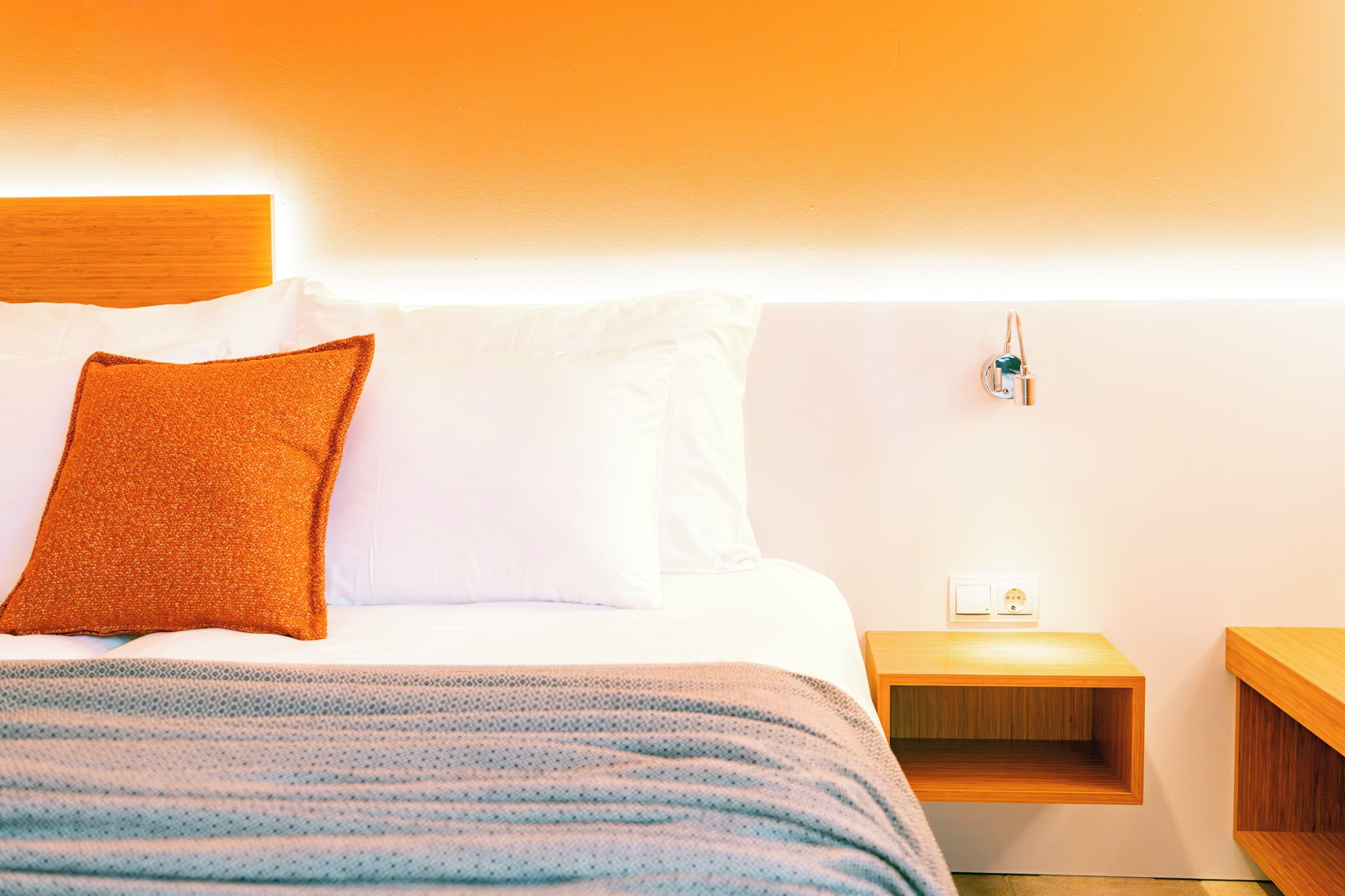 A neatly made bed with white and orange pillows, a blue and white striped blanket, and a wooden nightstand with a power outlet.