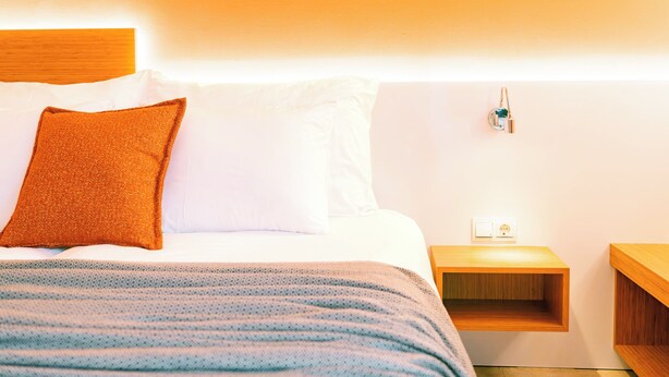 A neatly made bed with white and orange pillows, a blue and white striped blanket, and a wooden nightstand with a power outlet. - Calimera Sunpark Alanya (Alanya)