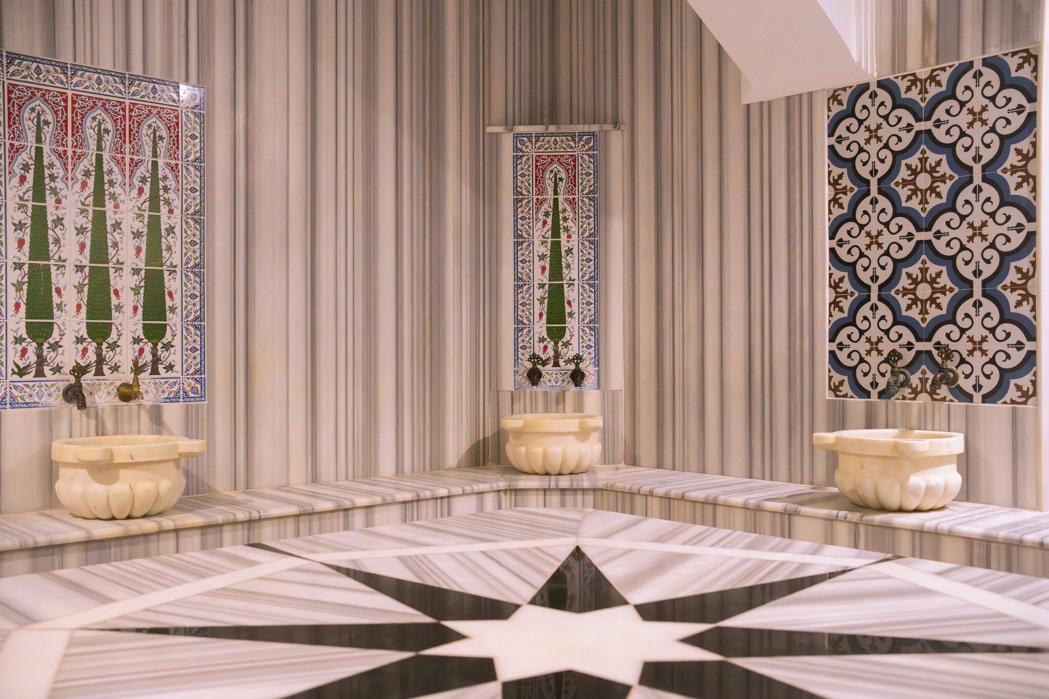 Une salle de bain avec des murs en marbre à rayures, trois vasques ornées de carreaux décoratifs et un sol à motifs étoilés.