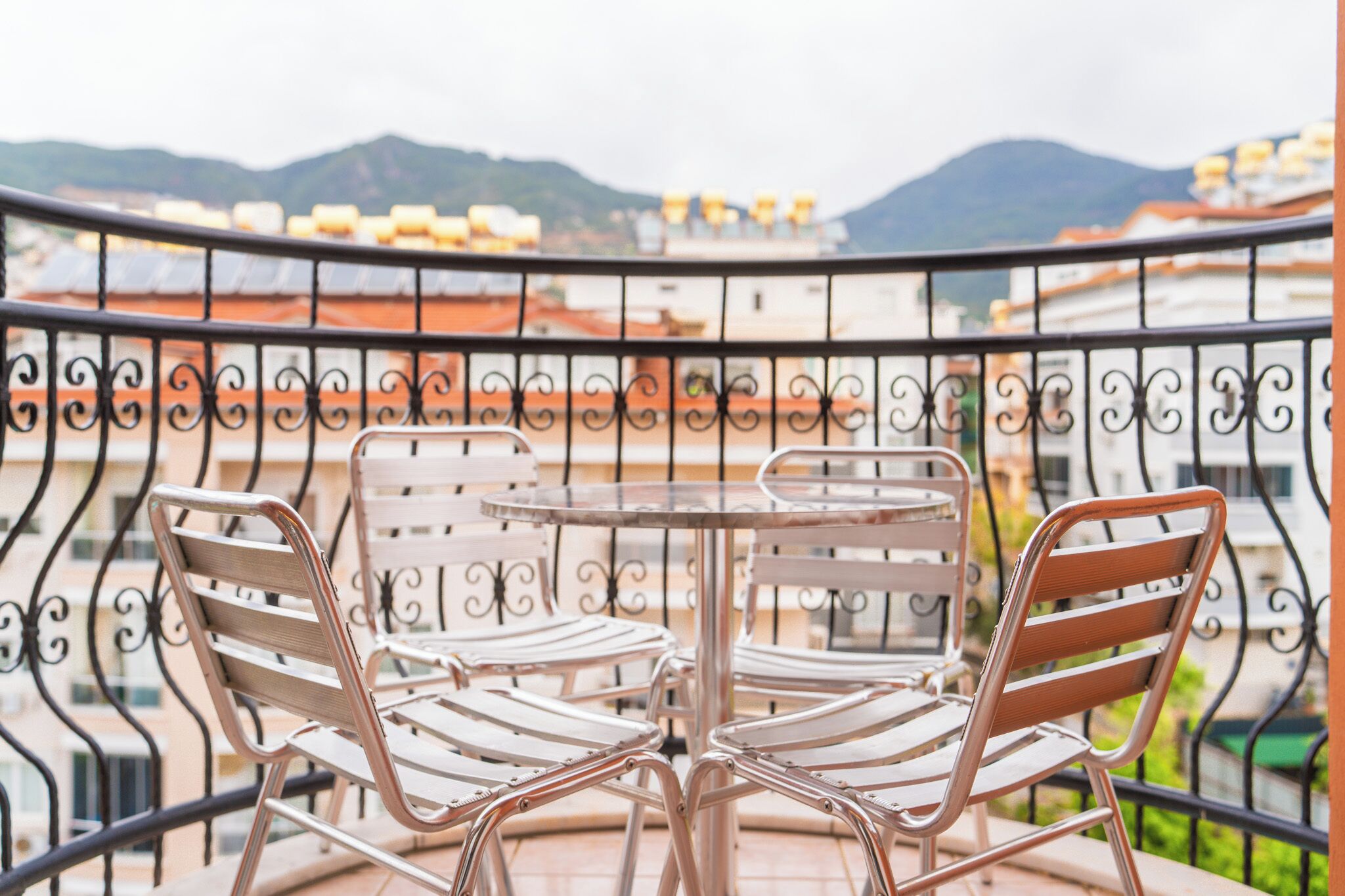 Ein Balkon mit rundem Tisch und vier Stühlen, mit Blick auf eine Stadtlandschaft mit Gebäuden und Bergen.