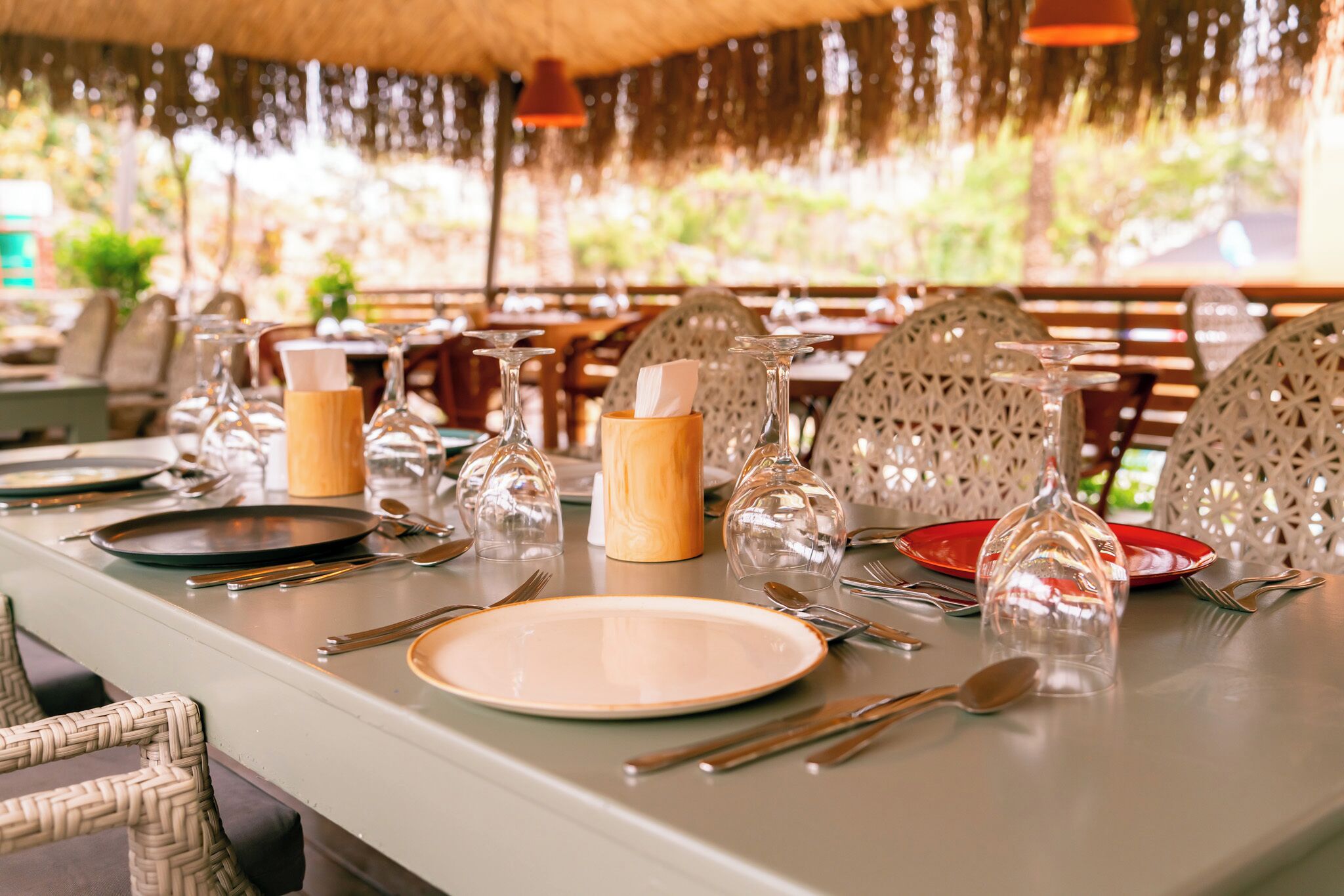 Mesa de comedor dispuesta con platos, cubiertos y cristalería, con sillas de mimbre y un techo de paja.
