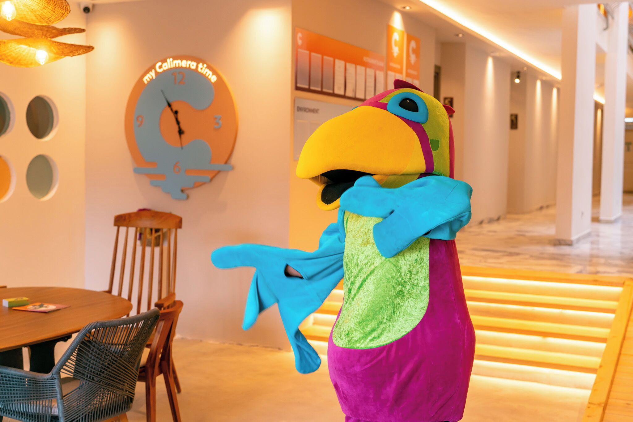 A hotel lobby with a large wall clock, a wooden dining table with chairs, and a colorful mascot.