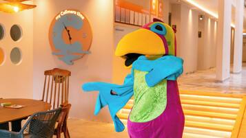 A hotel lobby with a large wall clock, a wooden dining table with chairs, and a colorful mascot.