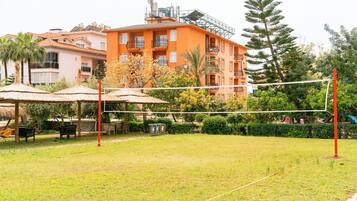 A grassy outdoor area with a volleyball net, umbrellas, and outdoor seating.