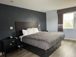 Um quarto moderno com cabeceira escura, uma cama com colcha cinza, uma mesa de cabeceira com um telefone e uma janela com cortinas.