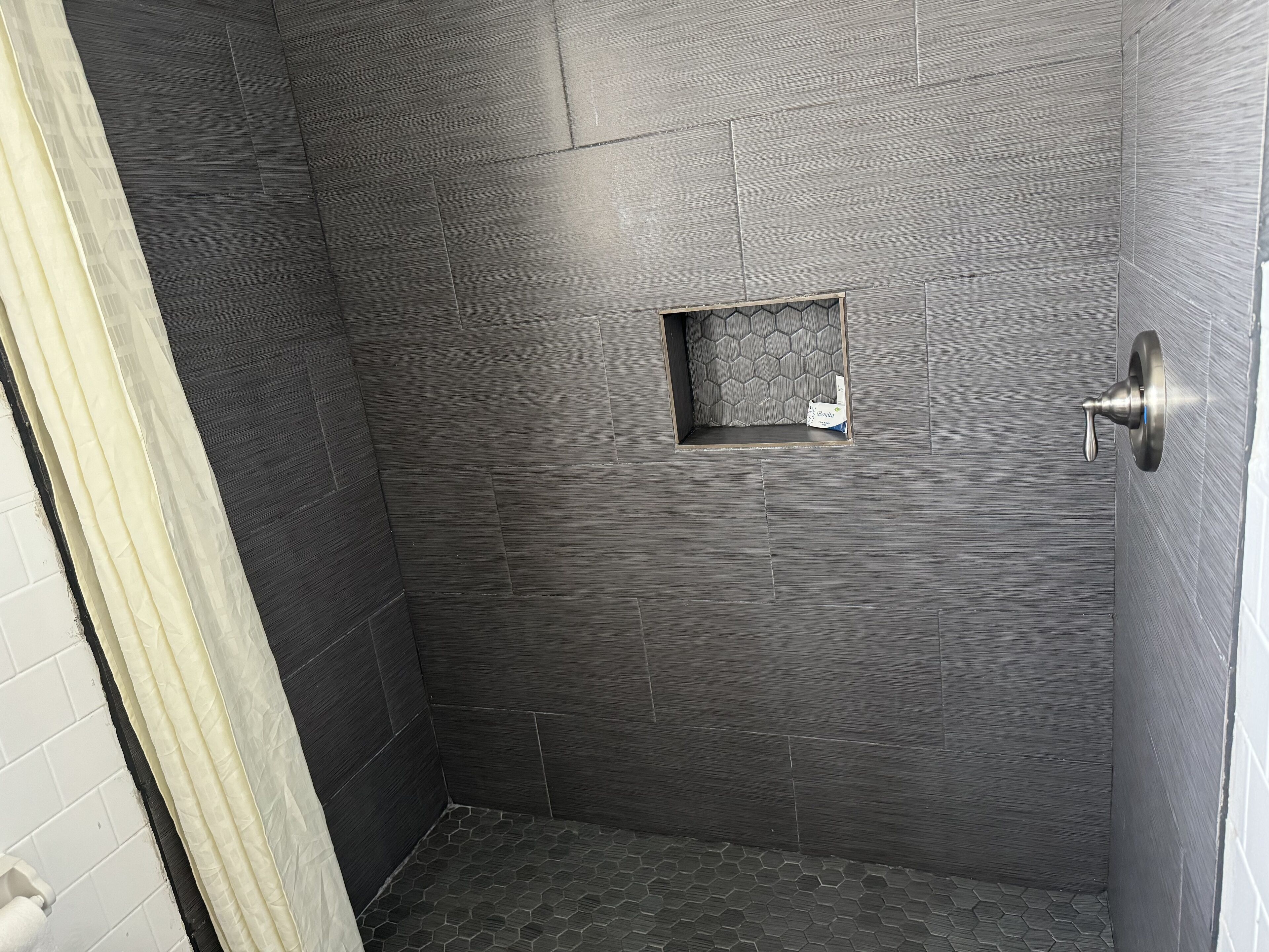 Une douche avec des carreaux sombres à texture, une petite fenêtre et un rideau de douche.