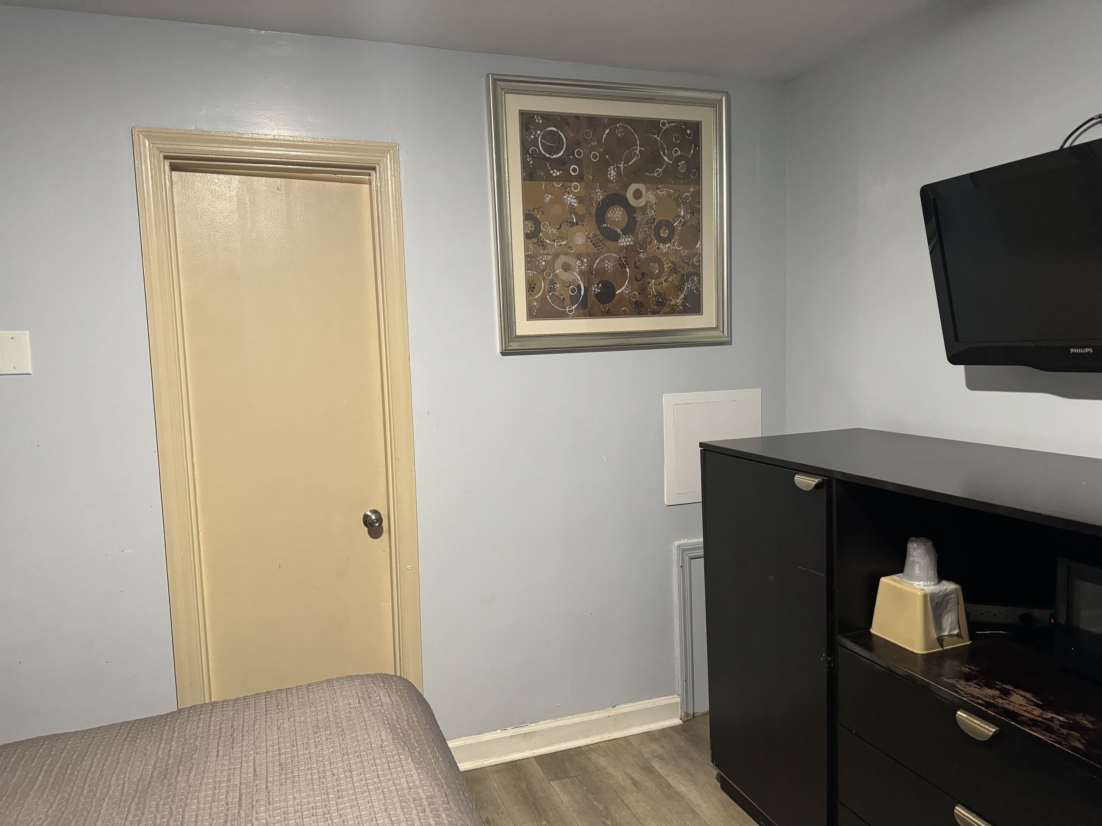 A room with a bed, a black TV on the wall, a framed abstract painting, and a black filing cabinet.
