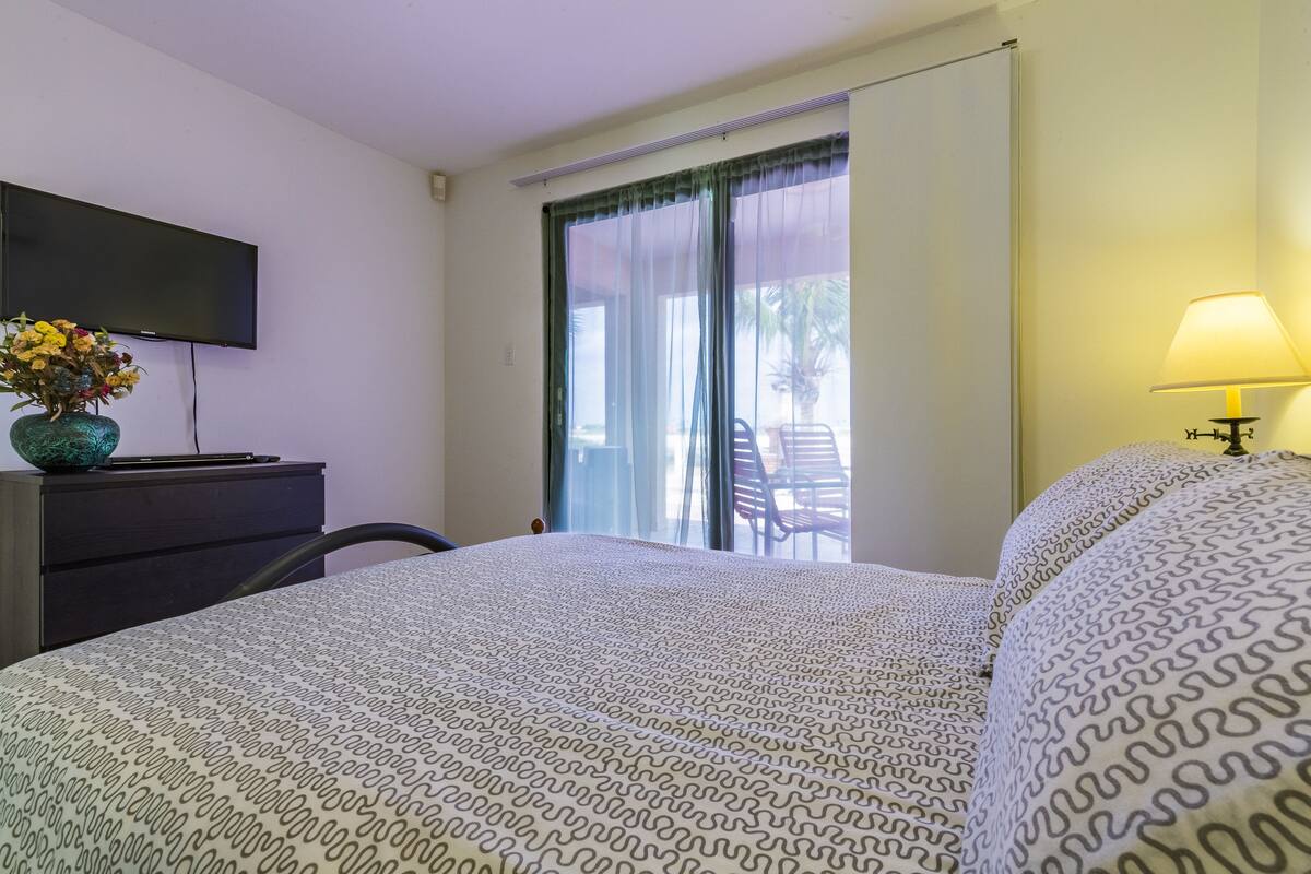 A hotel room with a bed, a desk with a flat-screen TV, a vase with flowers, a lamp, and a view of the outside balcony.
