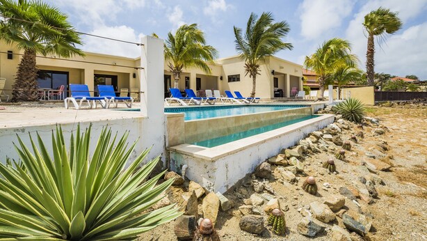 Un espace piscine aménagé avec des chaises longues, des palmiers et un bâtiment doté de nombreuses portes et fenêtres.
