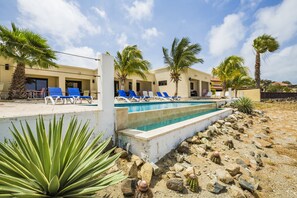 Un espace piscine aménagé avec des chaises longues, des palmiers et un bâtiment doté de nombreuses portes et fenêtres.