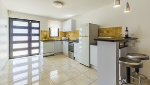 Une cuisine moderne avec des meubles blancs, une cuisinière et un réfrigérateur. On y trouve deux tabourets de bar et un petit îlot central sur lequel est posée une bouteille de vin.