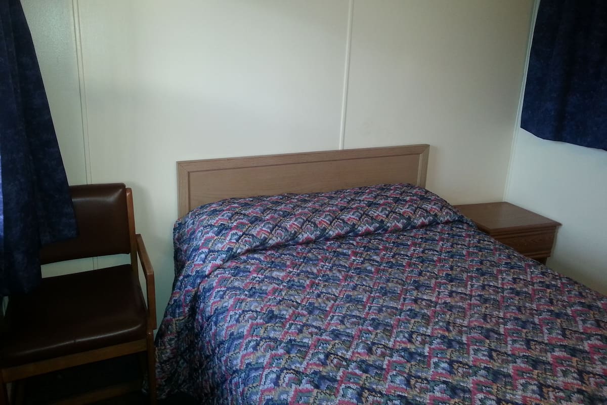 A single bed with a patterned bedspread, a wooden headboard, a wooden nightstand, and a brown leather chair.