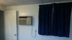A room with a white door, an air conditioning unit mounted on the wall, a dark curtain, and a wooden bench. - Americana Motel (Avenel)