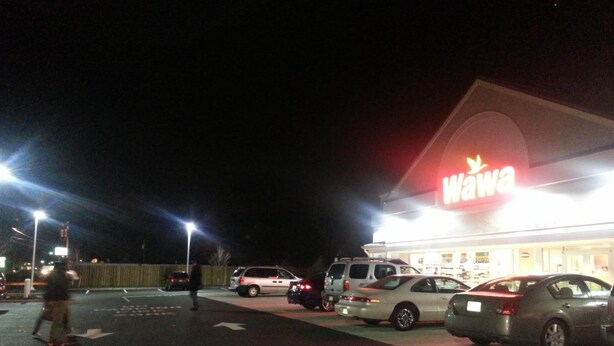 A parking lot with several cars and a pedestrian walking by a brightly lit store with a 'Wawa' sign. - Americana Motel (Avenel)