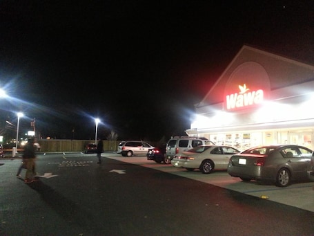 A parking lot with several cars and a pedestrian walking by a brightly lit store with a 'Wawa' sign.