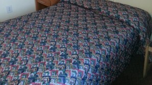 A room with a bed, a small wooden nightstand, a window with dark curtains, and a plain wall. - Americana Motel (Avenel)