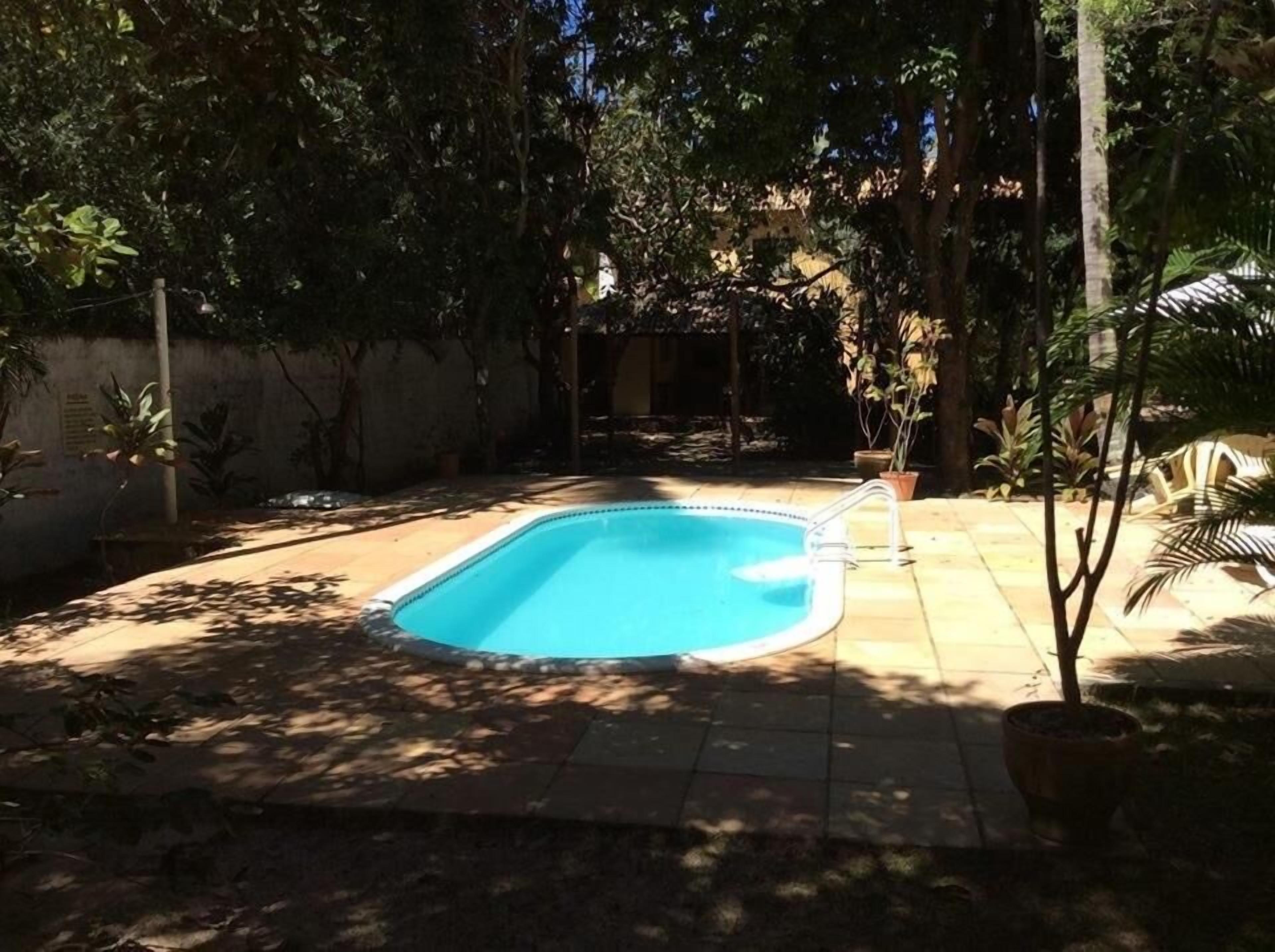 A rectangular swimming pool with a tiled deck surrounding it, set in a shaded backyard with trees and potted plants.