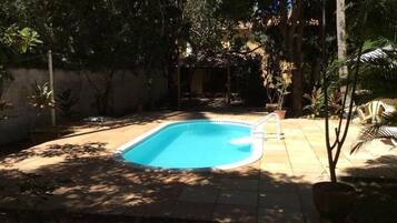 A rectangular swimming pool with a tiled deck surrounding it, set in a shaded backyard with trees and potted plants.