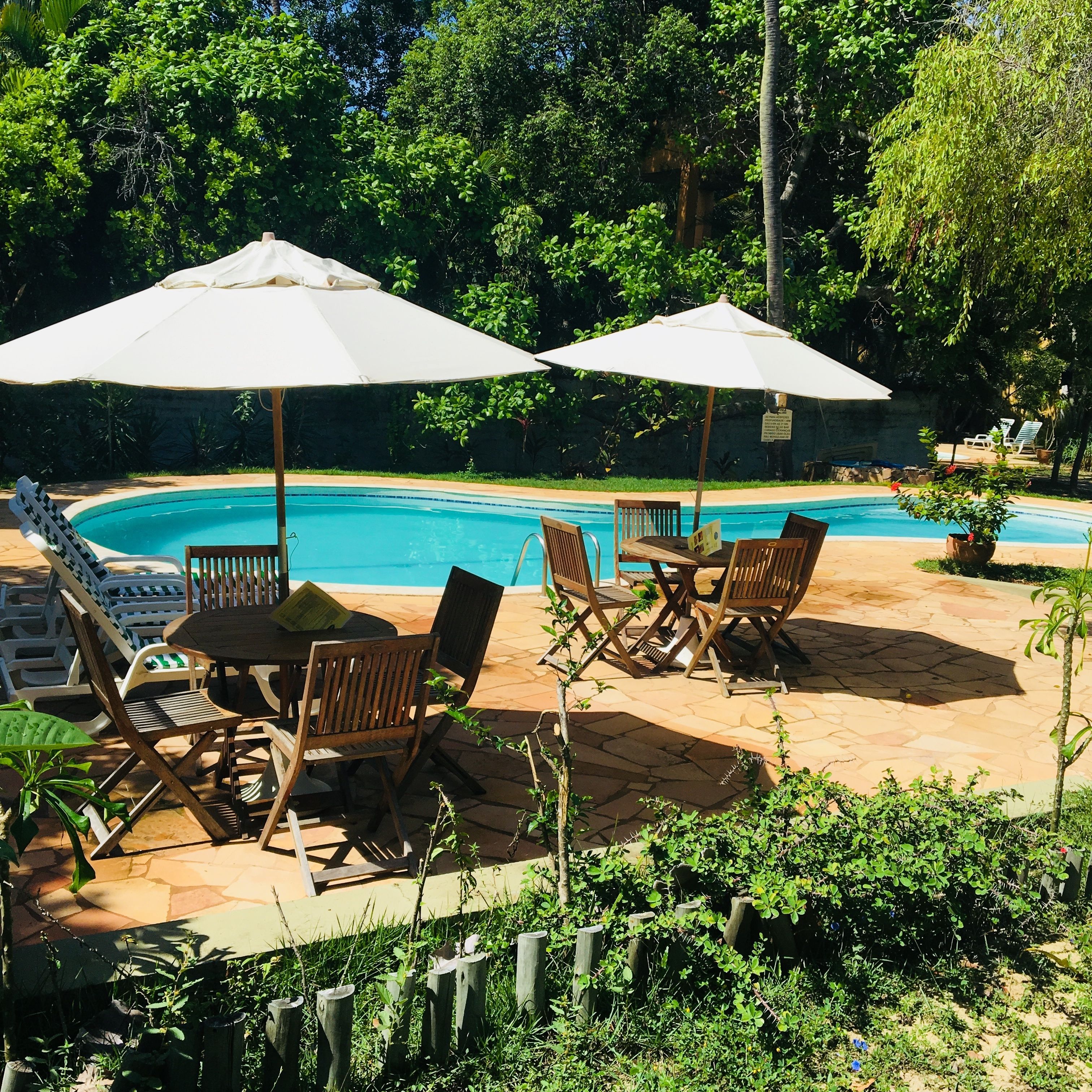 A poolside area with wooden chairs and tables, umbrellas, and a clear blue pool.