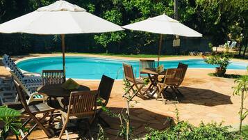 A poolside area with wooden chairs and tables, umbrellas, and a clear blue pool.