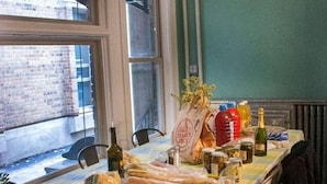 A dining area with a table set for six, covered with a checkered tablecloth. There are baguettes, bottles, and a bag labeled "Trader Joe's" on the table.