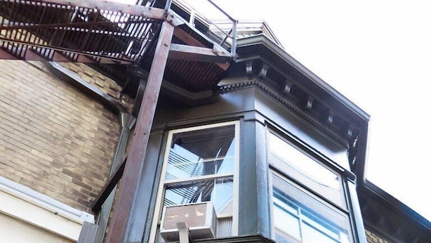 A corner of a building with a dark-colored exterior, a visible window, and a fire escape.