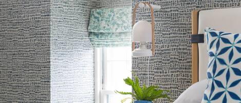 Un dormitorio con papel tapiz estampado, una cama con manta azul, una mesita de noche con una planta y una ventana con cortinas.